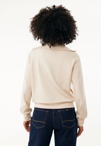 Sudadera beige con un cuello amplio y puños de canalé, combinada con jeans azul oscuro que presentan costuras decorativas y bolsillos traseros.