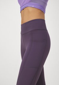Leggings morados con una cintura alta, que cuentan con un bolsillo lateral y detalles de textura perforada en una pierna. Tela suave y elástica.