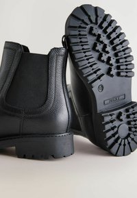 Botas Chelsea de cuero negro con suelas texturizadas, mostradas desde los lados y la parte trasera, destacando su diseño y material.