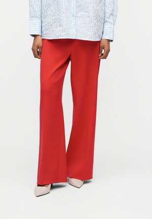 Personne portant un pantalon large rouge vif, une chemise bleue claire texturée et des talons hauts blancs à bout pointu, debout sur un fond blanc.