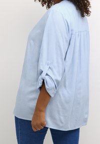 Frau mit dunkler Haut trägt eine hellblaue Langarmbluse mit hochgekrempelten Ärmeln und blaue Jeans, steht vor einem schlichten weißen Hintergrund.