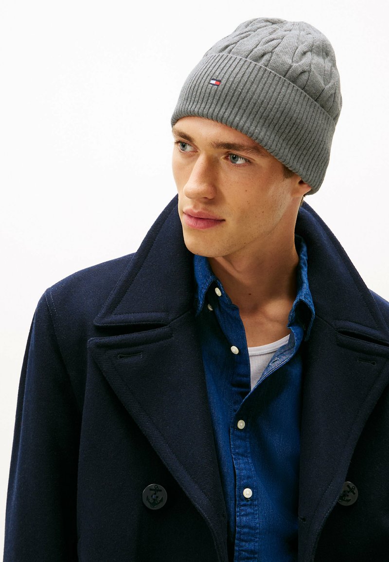 Gray knit beanie with cable pattern and ribbed edge, featuring a small logo. Worn with a dark navy coat and a blue shirt underneath.