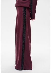 Bordowe spodnie o szerokich nogawkach wykonane z miękkiego materiału, z czarnym teksturowanym paskiem po boku. Prosty, luźny krój o gładkiej fakturze.