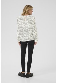 Chemisier blanc avec un motif ondulé noir, manches longues avec des poignets froncés, détail en trou de serrure au dos. Associé à un pantalon noir ajusté et des chaussures.