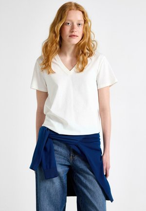 Rødhåret kvinde iført en hvid T-shirt med V-hals, blå jeans og en navyblå sweater bundet rundt om taljen mod en hvid baggrund.