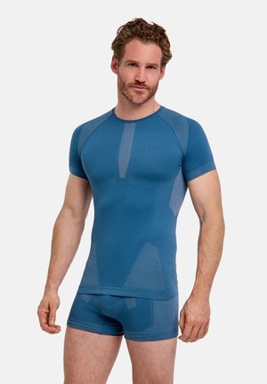 Man met kort krullend haar en baard, gekleed in een blauw aangesloten korte mouwen top en bijpassende korte broek tegen een effen lichte achtergrond.