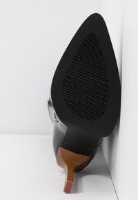 Chaussure à talon haut en cuir noir à bout pointu avec semelle en caoutchouc texturée et talon fin et incliné de couleur brune sur fond blanc.