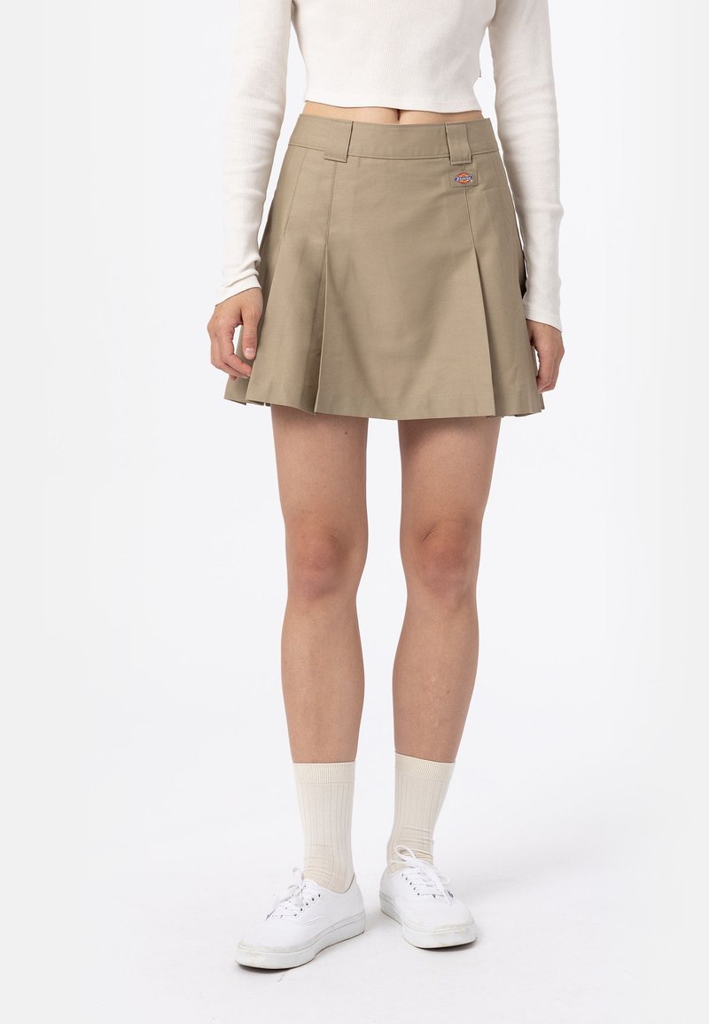 Femme portant une mini-jupe plissée beige, un haut blanc à manches longues, des chaussettes blanches et des baskets blanches, debout devant un fond uni.