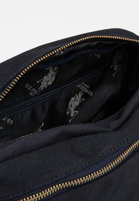 Laivastonsininen kangaslaukku kultaisella vetoketjulla, paljastaen sisäosan, joka on vuorattu tummalla kuviolla, jossa on vaaleanharmaita logokuvioita. Karkeapintainen ulkopuoli.