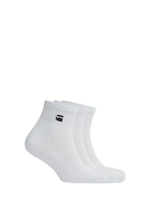 Trois paires de chaussettes blanches courtes avec bord-côtes et un petit logo noir sur le côté, disposées côte à côte.