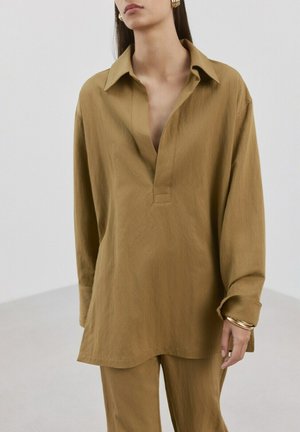 Femme portant une chemise ample à manches longues couleur moutarde avec col ouvert et pantalon assorti, accessoirisée de bracelets et boucles d'oreilles en or.