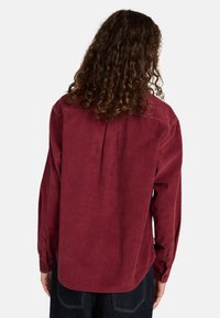 Rote cordartige Hemd mit langen Ärmeln, knöpfbaren Manschetten, einer Rückenfalte und einem abgerundeten Saum. Weiche Textur mit sichtbarem Rippmuster.
