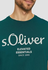 Tyrkysové bavlněné tričko s kulatým výstřihem, bílé nápisy "s.Oliver" a "ELEVATED ESSENTIALS SINCE 1969" v tučném písmu.