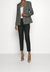 Kobieta w czarno-białej wzorzystej marynarce, białej bluzce, czarnych dopasowanych spodniach i srebrnych czółenkach na wysokim obcasie z paskami na kostkach.