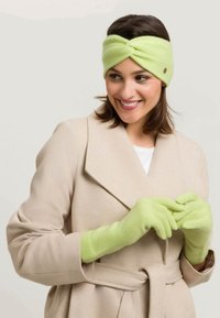 Bandeau tricoté vert clair avec design torsadé et gants assortis, porté avec un manteau beige. Texture lisse et détails discrets.