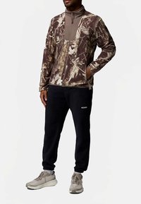 Kahevärviline kamuflaažiga fliisjakk pruunides ja beežides toonides, esiküljel tõmblukk; kombineeritud mustade dressipükstega ja heledate hallide spordijalatsitega.