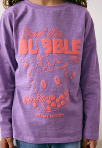 Lilla pika lõhikuga särk, millel on erksas korallitoonis multifilmiline bubble tea graafika, koos tekstiga "Magus nagu BUBBLE TEA" ja "ERILINE VERSIOON."