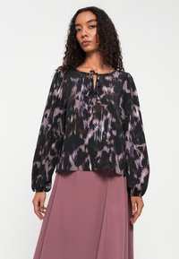 Schwarze Bluse mit lila und braunen abstrakten Mustern, langen Puffärmeln und einem Bindeband-Detail. Getragen mit einem rosa Rock.