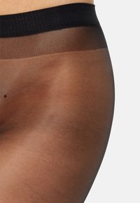 Gennemsigtige sorte strømpebukser med en bred sort taljeelastik, subtil glans og en glat tekstur, der delvist dækker et hudfarvet område.