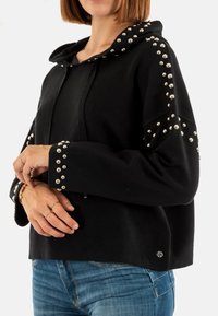 Sudadera negra con capucha, adornada con tachuelas plateadas a lo largo de los hombros y los puños. Tejido suave, diseño corto y corte holgado.