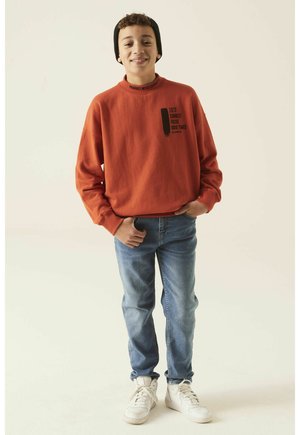 Felpa arancione con testo nero, jeans e sneakers bianche. La felpa ha un collo alto e una vestibilità comoda. Indossata con un berretto nero.