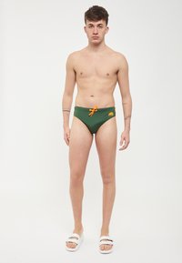 Grønne svømmedragt med en orange snor, der har et logo på fronten. Modellen står barfodet og har hvide slidesandaler på.