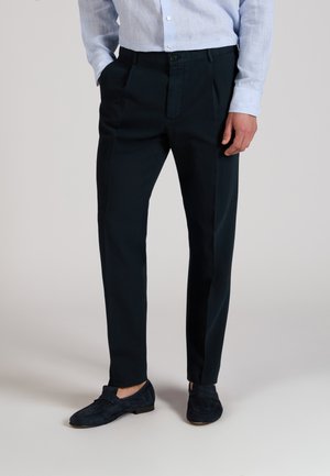 Hombre con pantalones negros a medida, camisa azul claro abotonada metida dentro, y mocasines de ante azul marino, de pie con una mano en el bolsillo.