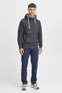 Grå trøje med hætte og kengurulomme, kontrasterende beige snoretræk og et logopatch, parret med marineblå bukser og grå sneakers.