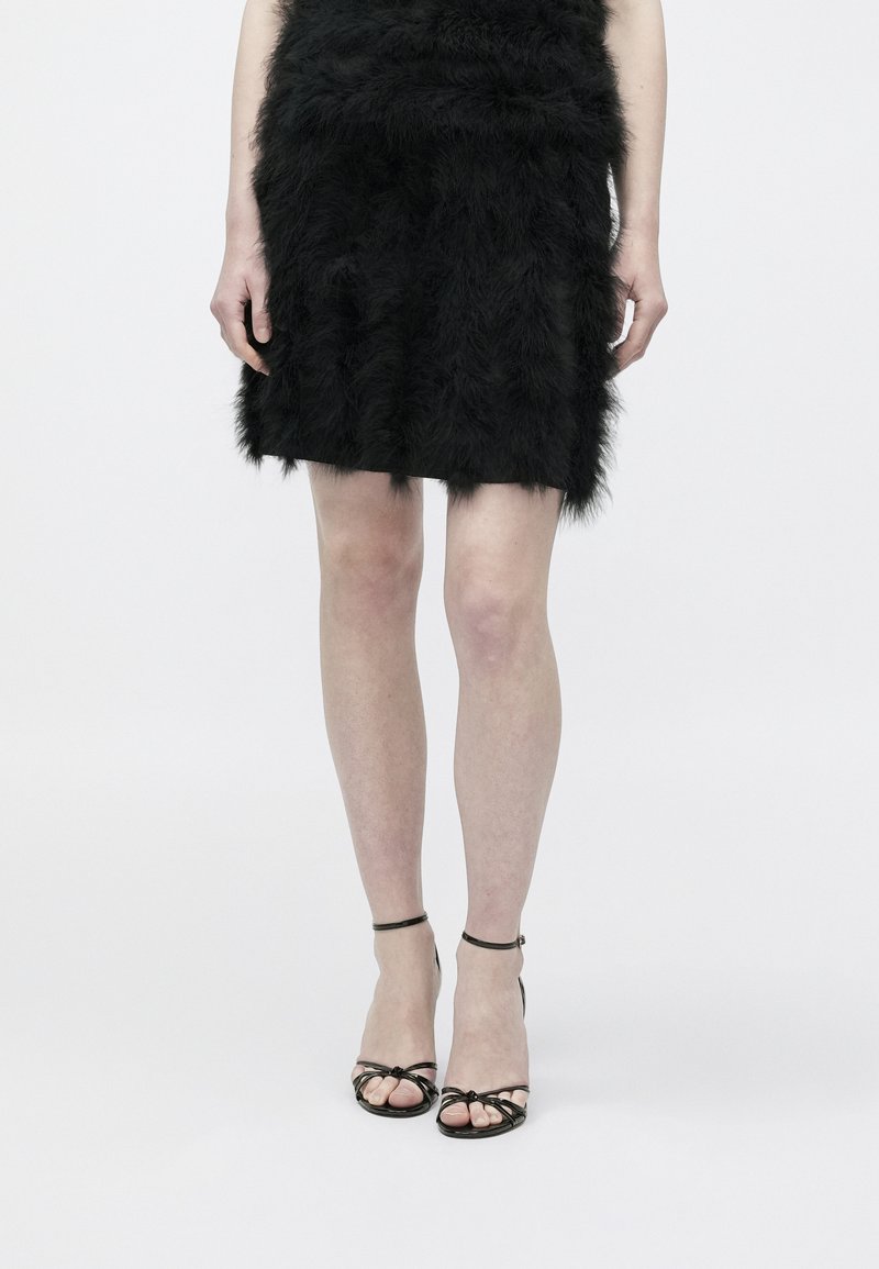 Μίνι φούστα από μαύρη συνθετική γούνα με στρωματοποιημένη υφή και ελαφριά γυαλάδα, συνδυασμένη με μαύρα σανδάλια με λουράκια και λεπτή διακοσμητική φιόγκο.
