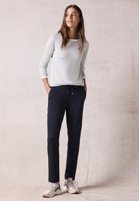 Grijze sweatshirt met lange mouwen en een ronde halslijn, gecombineerd met marineblauwe broek met trekkoord. Model draagt witte en grijze sneakers.