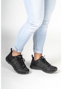 Svarta atletiska skor med övre del i texturerad stickad väv och svart gummisula. Har snörningar, dragögla och vadderad sula. Bärs med ljusblå jeans.