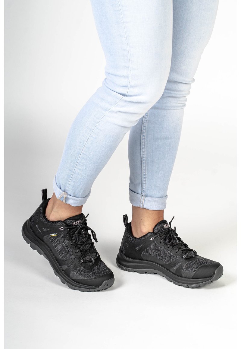 Svarta atletiska skor med övre del i texturerad stickad väv och svart gummisula. Har snörningar, dragögla och vadderad sula. Bärs med ljusblå jeans.