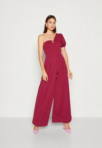 Macacão bordô de um ombro só com corpete estruturado, manga bufante, pernas largas e pregas; combinado com saltos altos cor-de-rosa.