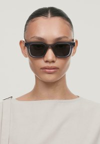 Occhiali da sole neri oversize con una montatura grigia trasparente e spessa. Lenti lisce e forma rettangolare. Il modello indossa un top di colore chiaro.