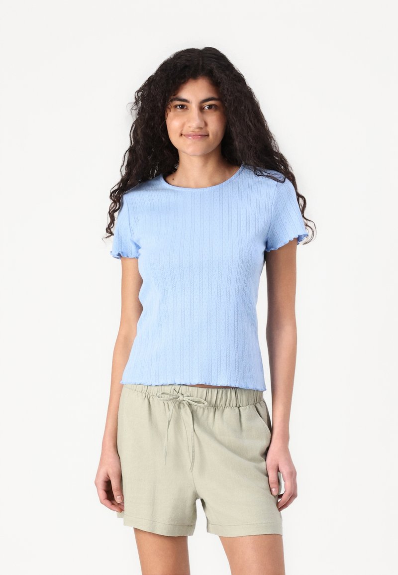 Lyseblå strikket kortermet topp med ribbestruktur og bølget kant, kombinert med beige shorts med snøring.