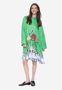 Vestido camisa de satén verde con patrones florales y geométricos, con cuello, mangas largas y un dobladillo con un borde triangular en blanco y negro.