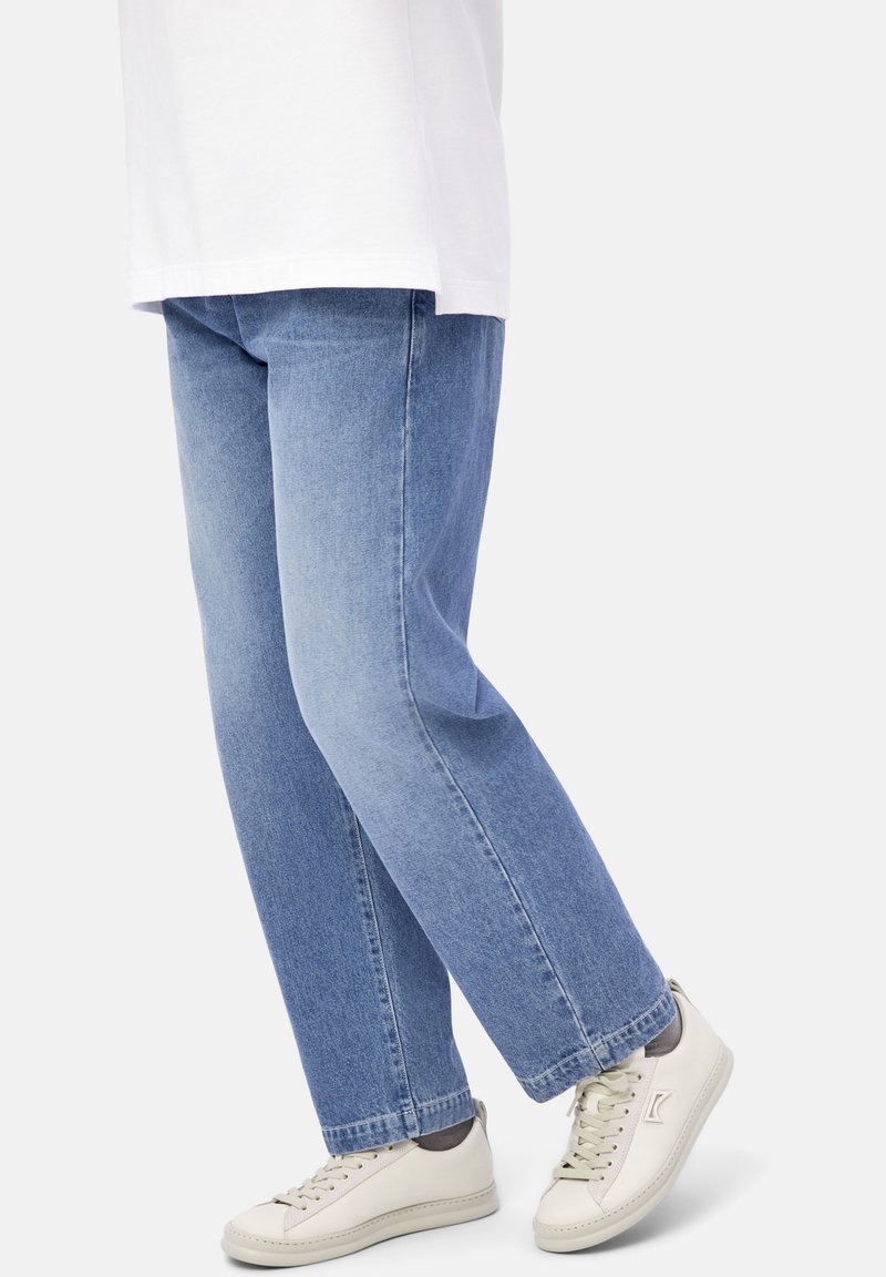 Person wearing light blue jeans and white sneakers with gray socks, standing with legs crossed against a plain background.