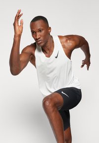 Weißes sportliches Tanktop mit Netzstruktur und schwarzen Akzenten; schwarze Shorts mit einem wei�ßen Logo. Modell in Laufpose.
