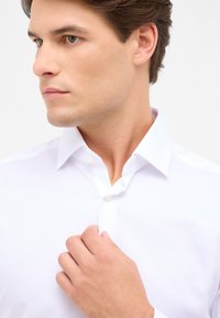 Witte button-up shirt met een gestructureerd patroon, voorzien van een puntige kraag en een enkele knop op de sluiting. Model dat de kraag aanpast.