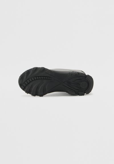 Black rubber shoe sole with distinct traction pattern, featuring grooves and spikes for grip. Smooth surface with slight curvature.