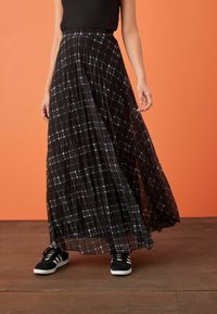 Sort ternet maxi-nederdel med en gennemsigtig tekstur, plisseret design og kontrasterende hvide linjer; parret med sort og hvide sneakers.