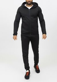 Survêtement de sport noir avec hoodie zippé, présentant un design ajusté et des poches. Associé à des chaussures noires avec des lacets orange.