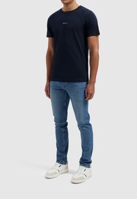 Marineblauwe katoenen T-shirt met een ronde halslijn en korte mouwen, met een witte geprinte logo op de voorkant. Gedirigeerd met lichtblauwe jeans.