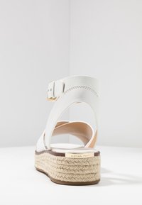 Valkoinen nahkainen espadrille-mallin korkokenkä, jossa on nilkkanyöri, kudottu jutee-alusta ja kultasävyiset yksityiskohdat. Pehmeä vuori ja ompeluyksityiskohdat.