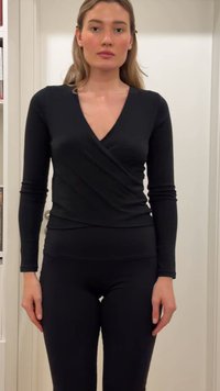 Femme debout à l'intérieur portant un haut cache-cœur noir ajusté à manches longues et des leggings noirs, devant un fond blanc avec une étagère.