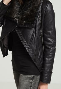 Veste en cuir noir avec col en fausse fourrure, fermeture éclair asymétrique et accents métalliques. Finition texturée avec détails à ourlet effiloché.