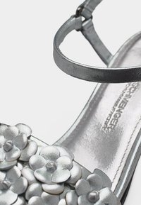 Silberne Sandale mit Riemen um den Knöchel und dekorativen metallischen Blumenapplikationen im Zehenbereich.