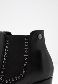 Stivale alla caviglia nero con esterno in pelle, pannello elastico e dettagli in metallo argentato lungo il lato, completato da un pulsante con logo vicino alla parte superiore.