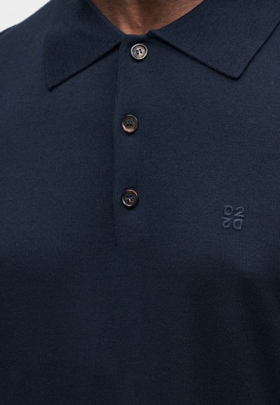 Μπλε ναυτικό πόλο μπλουζάκι από υφή ύφασμα με γιακά, διαθέτει τρία καφέ κουμπιά και ένα μικρό κεντημένο λογότυπο στο στήθος.
