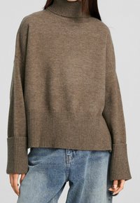 Brązowy, oversize sweter z golfem wykonany z miękkiego materiału dzianinowego z prążkowanymi mankietami i dołem. W połączeniu z jasnoniebieskimi, luźnymi dżinsami.